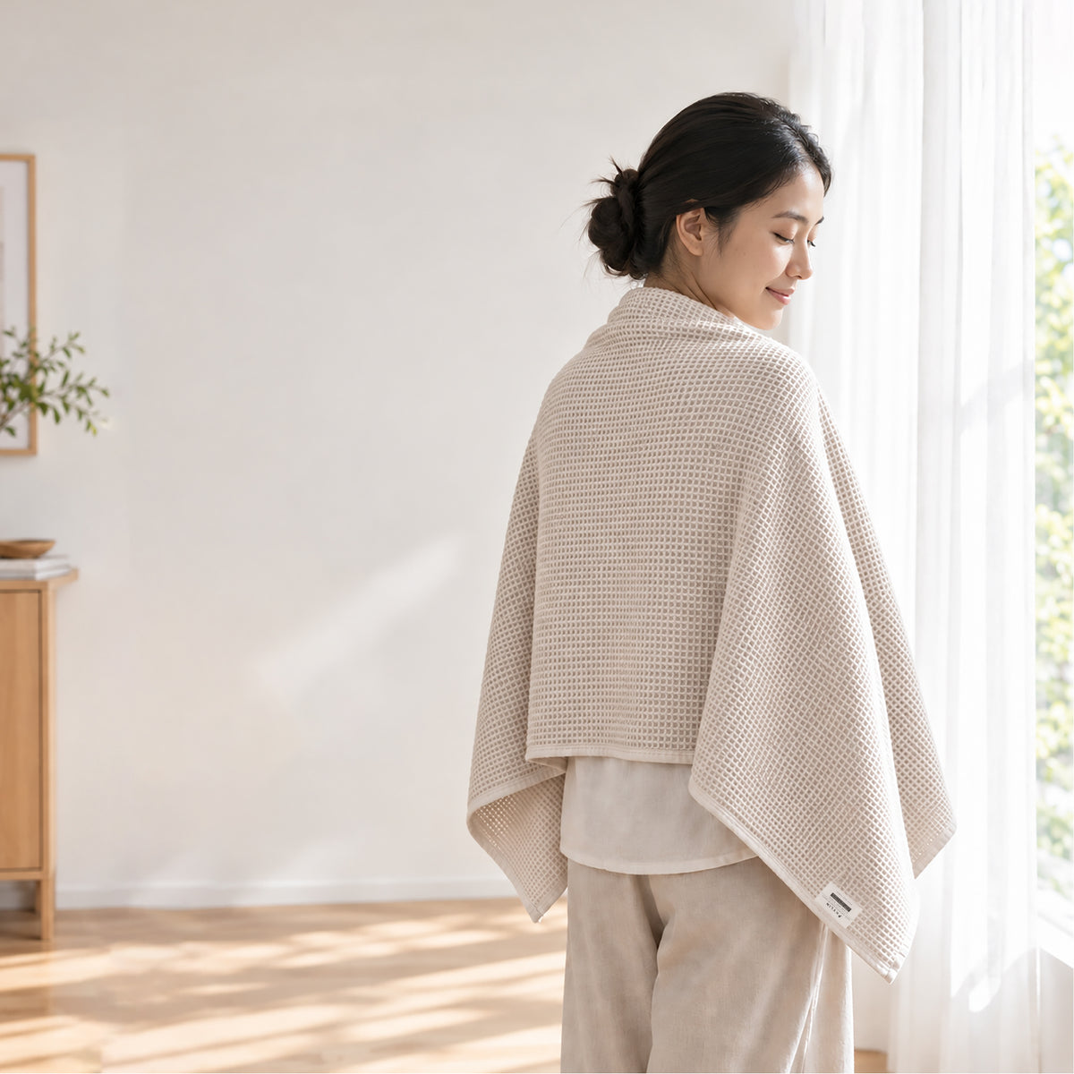 A serene woman wrapped in the cream Kylin Wellness honeycomb towel by a sunlit window, demonstrating how natural ambient light effortlessly activates the LumiPur self-purifying technology.