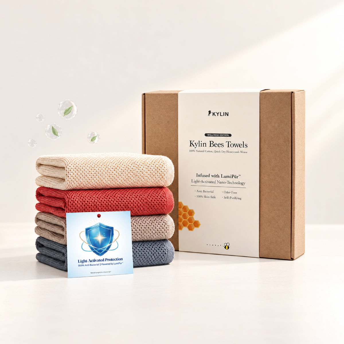 A set of folded Kylin bees towels in beige, red, brown, and slate blue, displayed next to their LumiPür™ brown cardboard packaging box and a blue informational LumiPür™ antimicrobial card on a clean white background.